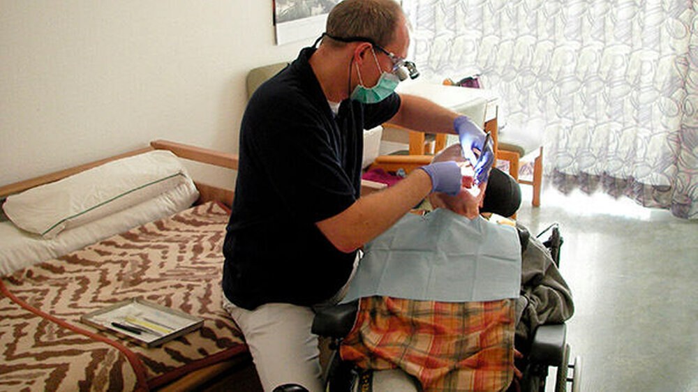 Der Patient sitzt im Pflegerollstuhl: Bei Pflegerollstühlen kann ohne großen Aufwand die gesamte Rückenlehne nach hinten gekippt werden (bei Unsicherheit hilft das Pflegepersonal). Als Behandler dann etwas erhöht zu sitzen, etwa auf dem höhenverstell