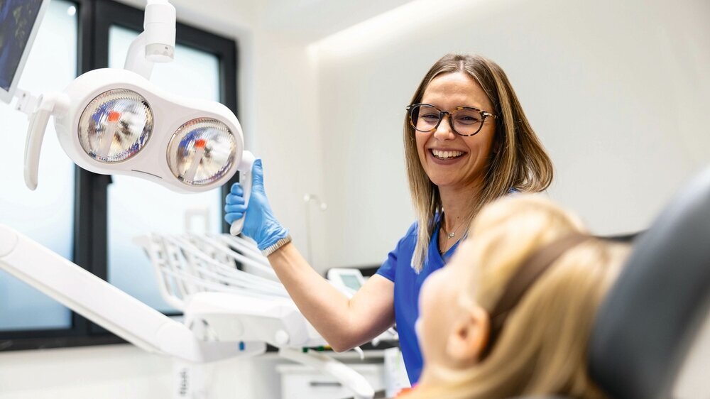 A dentist wearing glasses and gloves interacts with her young patient in a modern and brightly lit dental clinic, emphasizing care and professionalism.