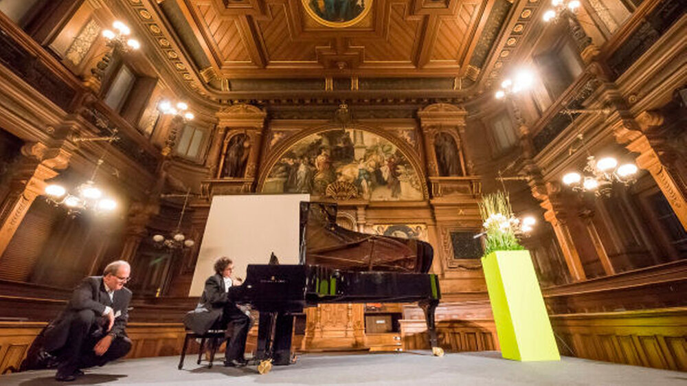 Alberto spielt erstmals vor Publikum mit der Beißschiene am Flügel der alten Aula der Universität Heidelberg. Im Vordergrund Entwickler Dr. Rüdiger Rupp, der gespannt Motor und Pedal beobachtet.