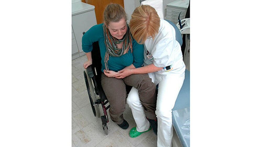 Abb. 10: Bei Hemiplegie, in der vorliegenden Situation linksseitig, wird der Rollstuhl so positioniert, dass die gelähmte Körperseite Kontakt zum Behandlungsstuhl hat. Die betreuende Person setzt sich auf den Behandlungsstuhl und legt das gelähmte Bein der Patientin über den eigenen Oberschenkel. Der gelähmte Arm wird mit der einen Hand stabilisiert. Die andere Hand umfasst die Patientin. Nun kann durch kleine Rutschbewegungen der Transfer auf den Behandlungsstuhl erfolgen. Die Patientin unterstützt die Bewegung – soweit möglich – mit dem gesunden Arm. |