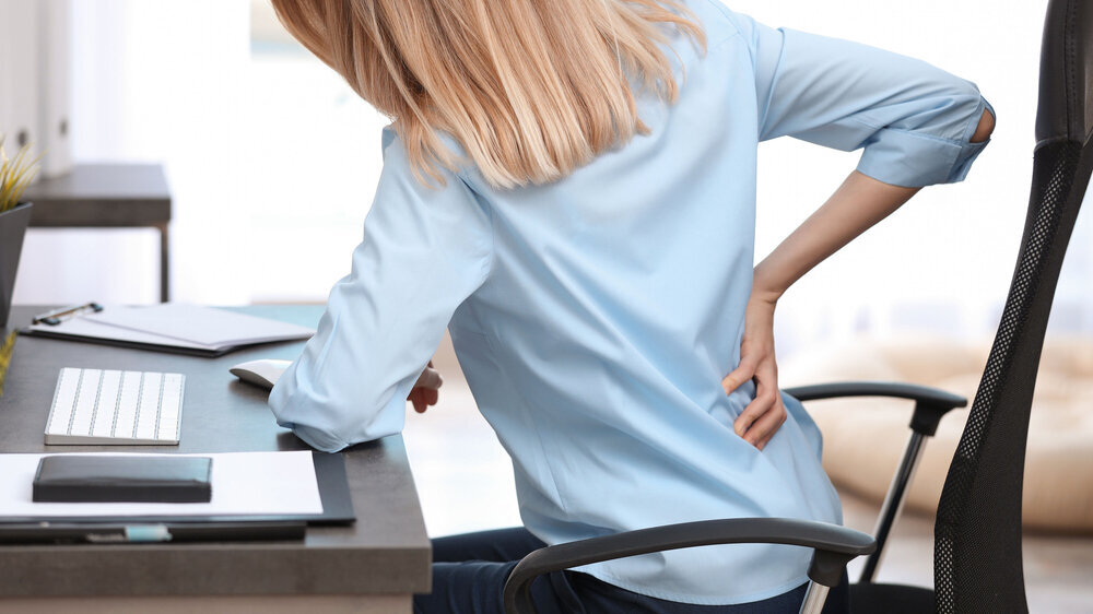 Young woman suffering from back pain in office Der TK-Gesundheitsreport 2023 wertet die Krankschreibungen und Arzneiverordnungen der rund 5,6 Millionen bei der TK versicherten Erwerbstätigen aus.